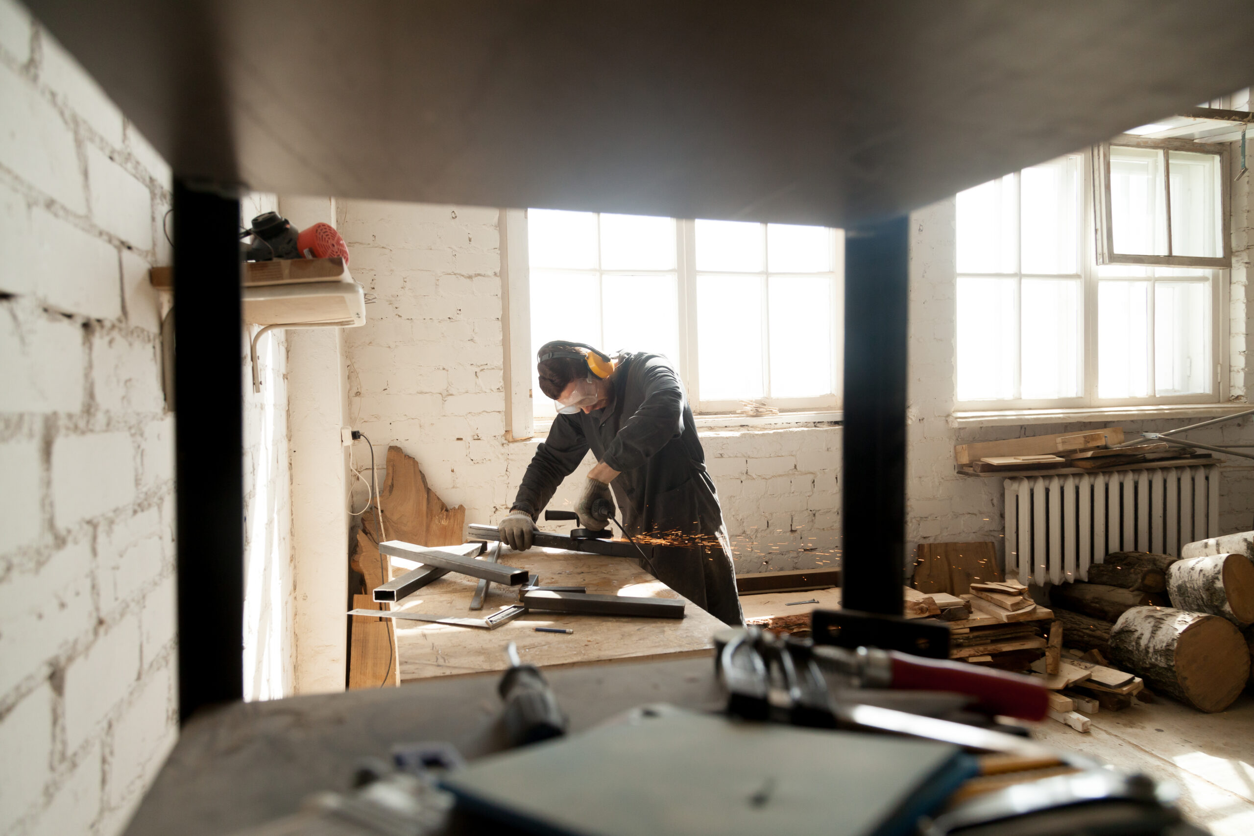 Trained young workman grinding metal parts of construction on table in workshop interior with tools equipment. Manual worker wearing protective clothes working with angle grinder in manufactory